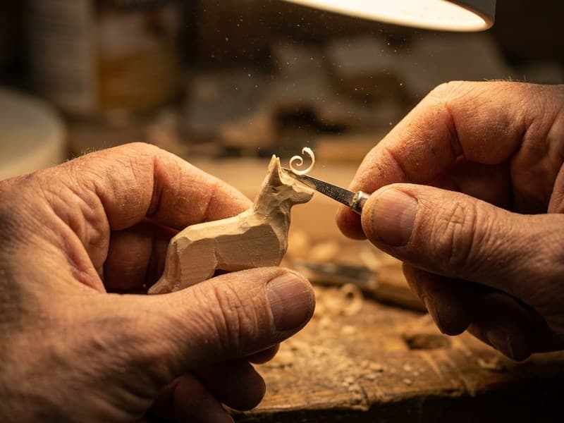 Close-up of hands using a detail knife to carve the ear of a miniature Belgian Malinois from basswood, with wood shavings curling away