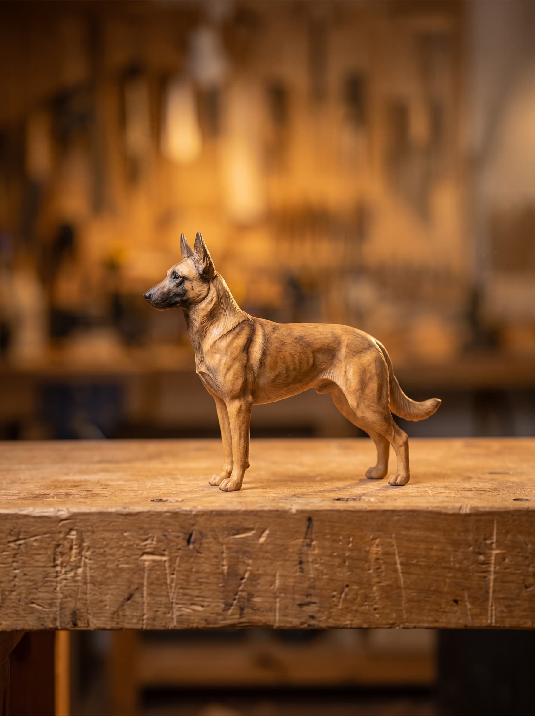 Belgian Malinois sculpture standing in full profile on a worn workbench, tan coat with a dark mask and carved muscle definition, tool wall softly lit behind it